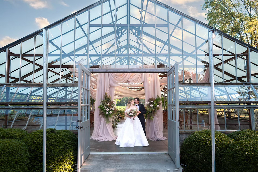 Bride poses in elegant gown at glass greenhouse wedding venue entrance with pink draping and floral arrangements.