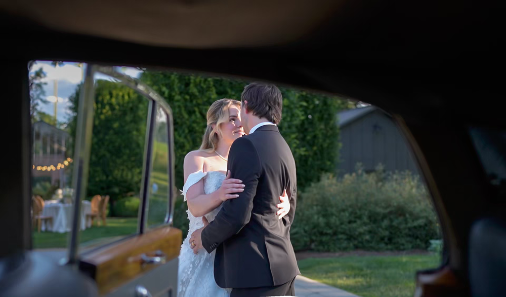 Bride and groom share a romantic moment beside a luxury car at an outdoor wedding venue.