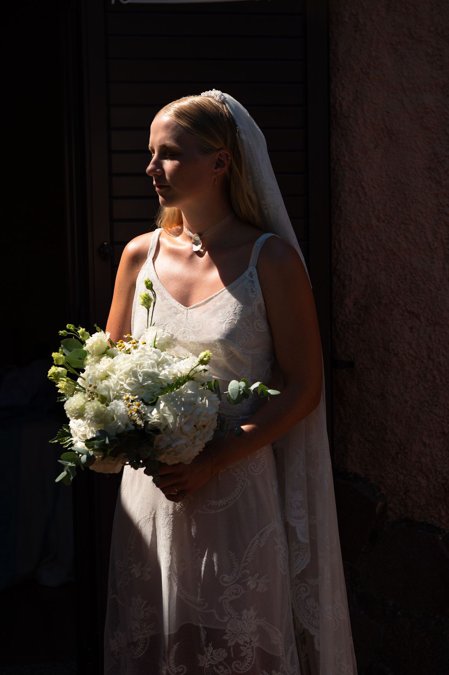 Bride in white lace gown and veil holds white floral bouquet, standing in dramatic sunlight against dark background.