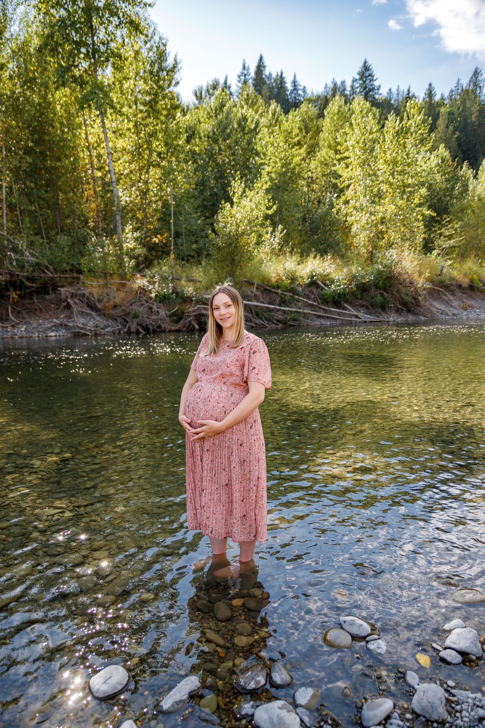 Pregnant woman in pink floral dress standing in a rocky river surrounded by lush green trees on a sunny day.