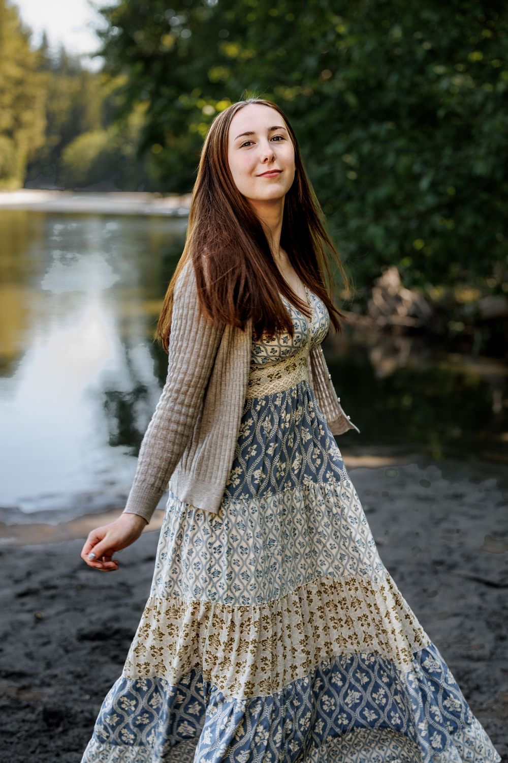 Young woman in floral maxi dress and cardigan standing by a serene river surrounded by lush green trees.