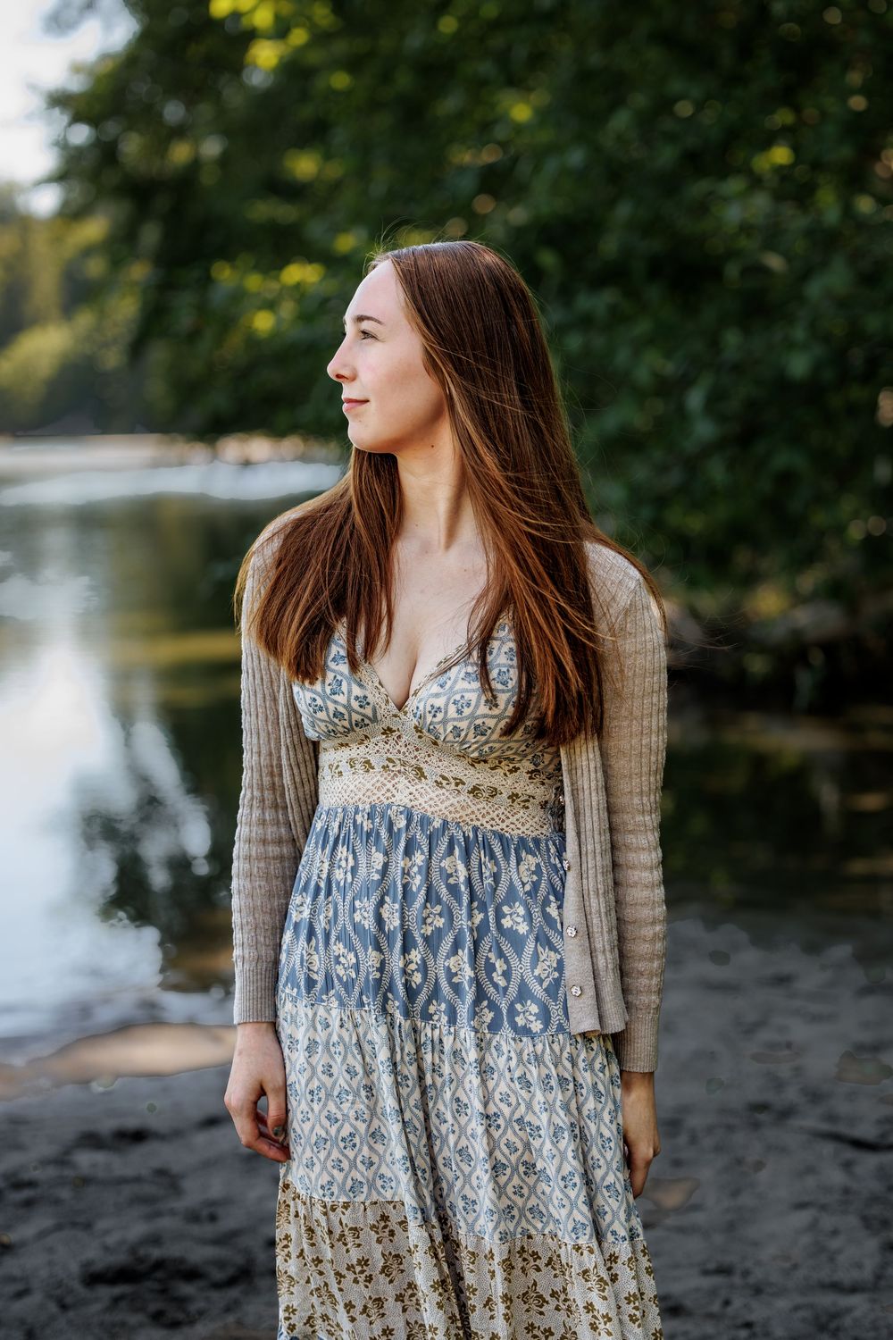 Young woman in floral maxi dress and cardigan standing by a serene river surrounded by lush green trees.