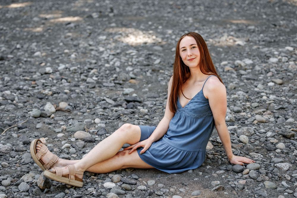 Young woman with long red hair sits on rocky ground wearing a blue sundress and tan sandals, smiling outdoors.