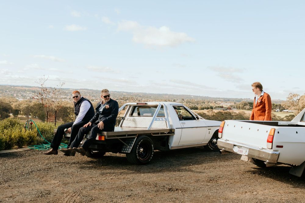 Three groomsmen relax against a white flatbed ute truck in a rural landscape under a blue sky.
