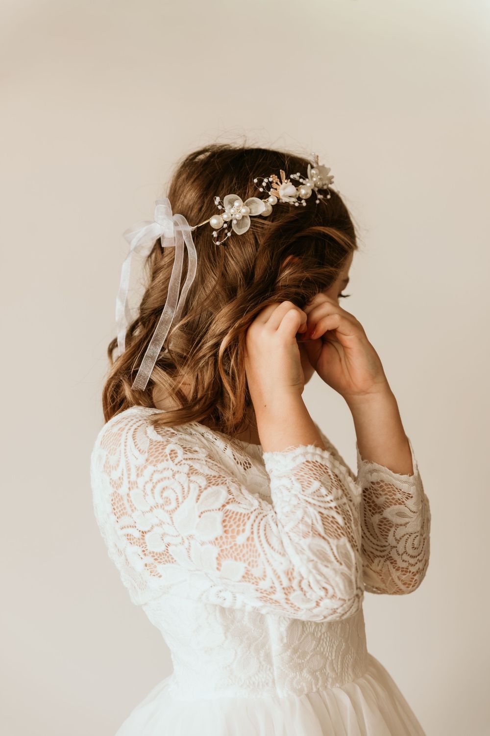 A bride in a lace long-sleeve wedding dress adjusts her floral hair crown from behind.