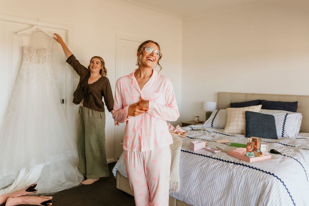 A bride in pink pajamas holds a drink while a bridesmaid displays her lace wedding dress in a bedroom.