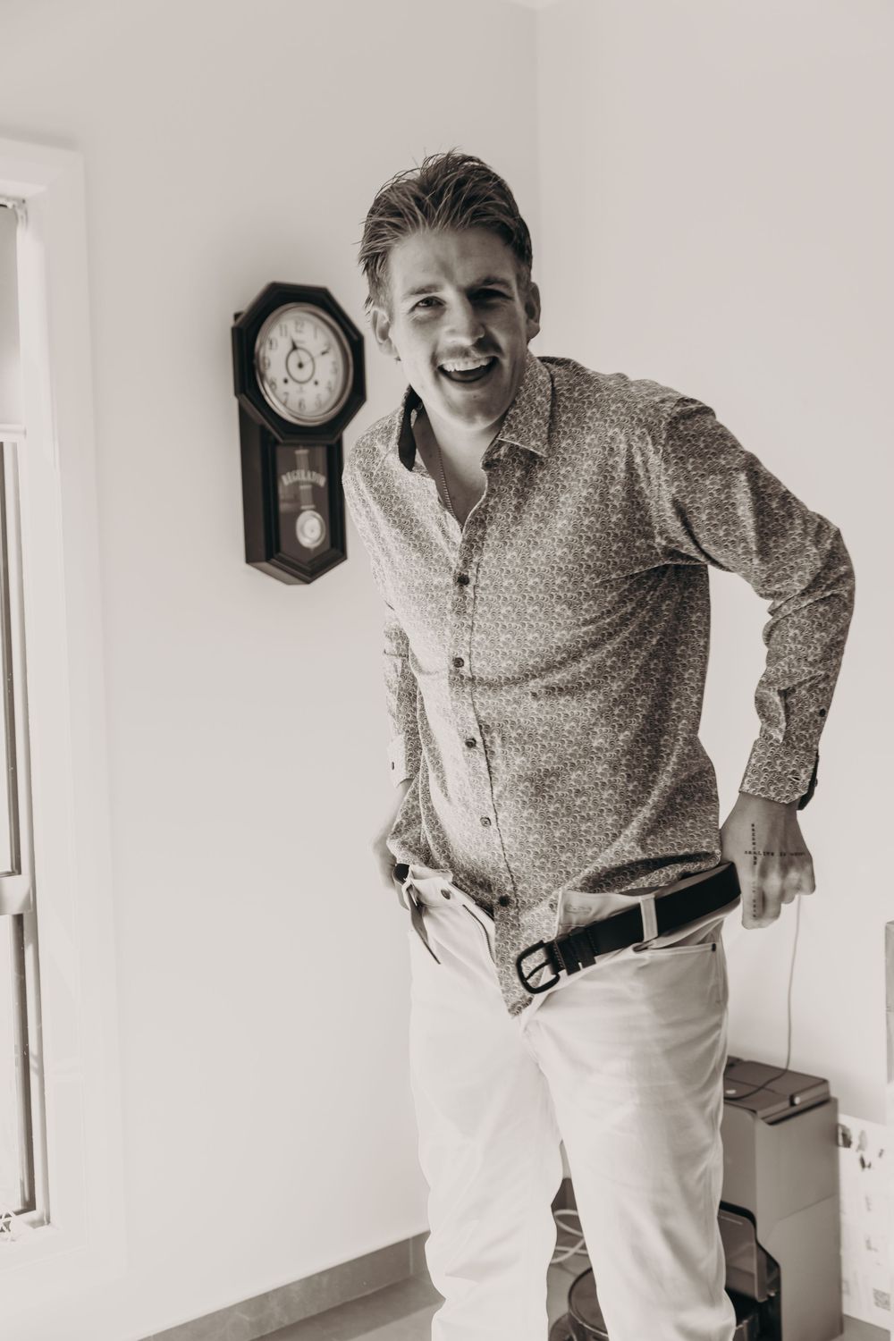 A laughing young man in a patterned shirt and white pants poses beside a vintage wall clock indoors.