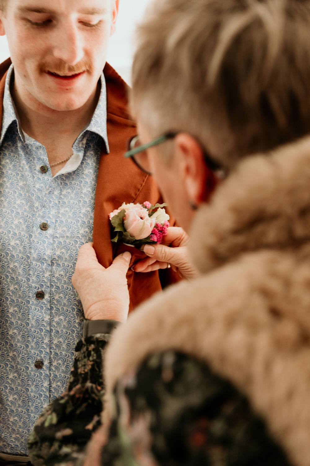 An older woman pins a pink boutonniere onto a groom's rust-colored jacket before the wedding ceremony.