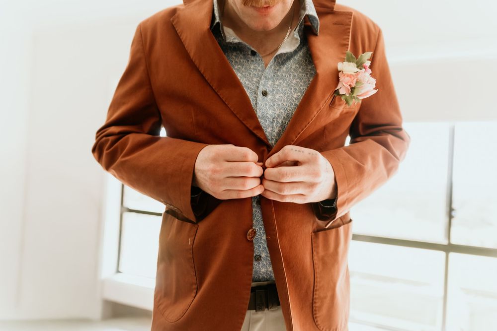 A groom in a rust-brown suit jacket buttons his coat with a pink floral boutonniere on his lapel.