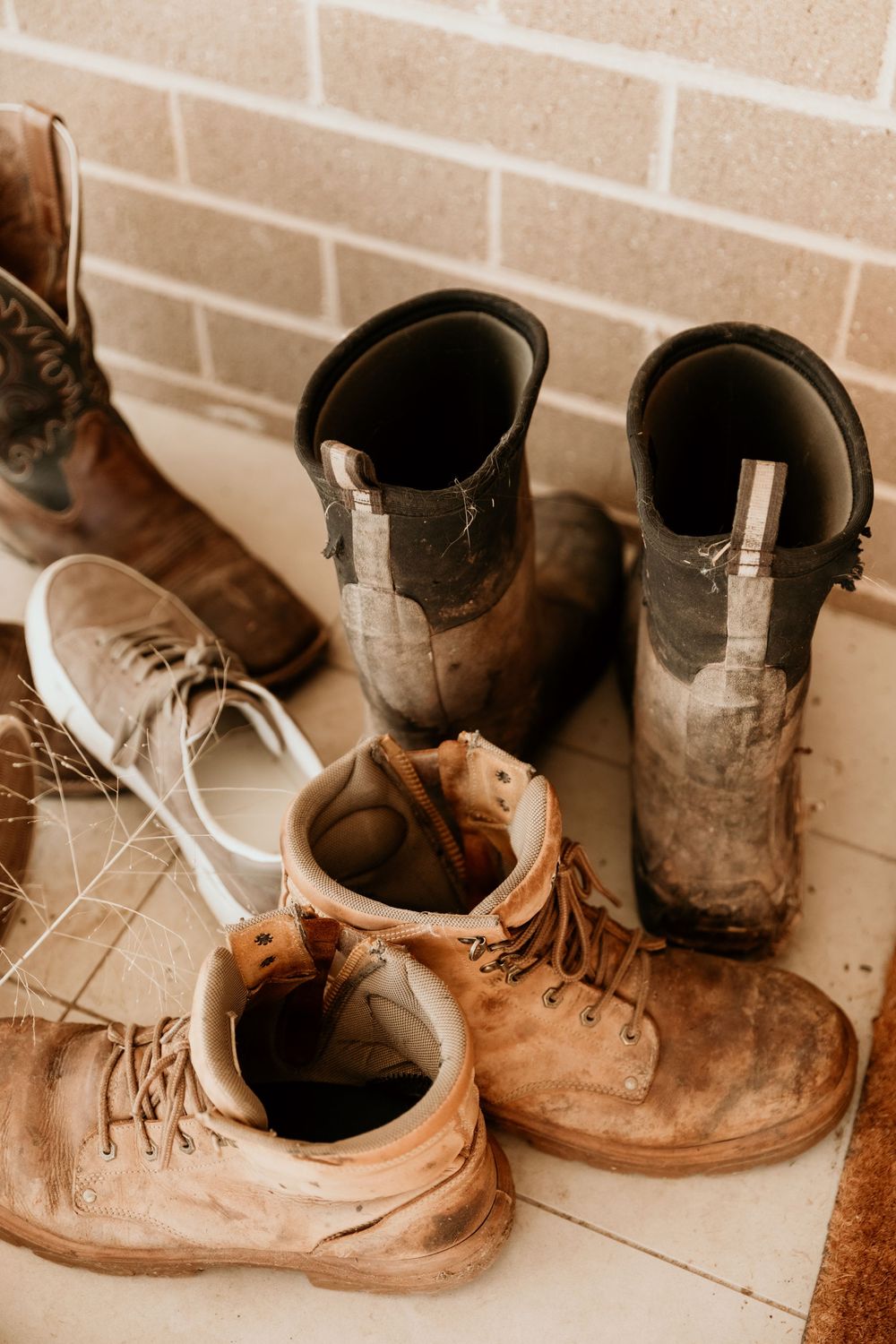 A collection of worn muddy boots and shoes arranged on a tiled floor against a brick wall.
