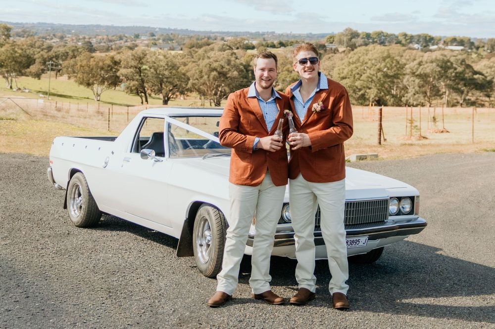 Two smiling men in matching brown jackets hold beer bottles while leaning on a classic white car outdoors.