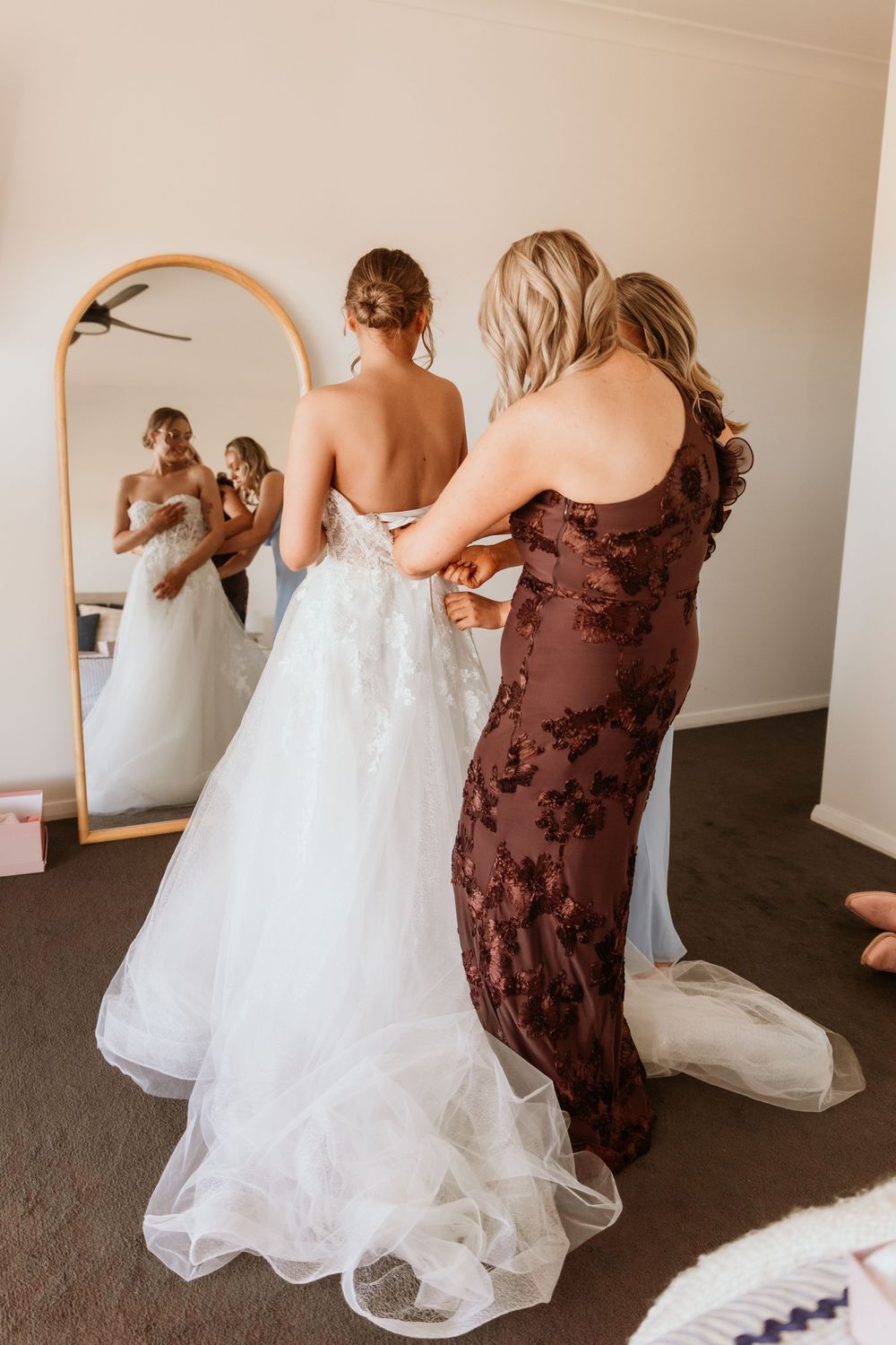 A bridesmaid in a dark floral gown laces up the back of a bride's white tulle wedding dress.