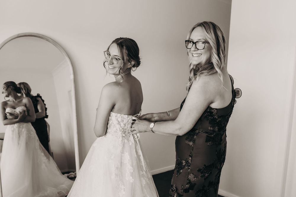 A black and white photo of a woman fastening the back of a bride's wedding dress near a mirror.