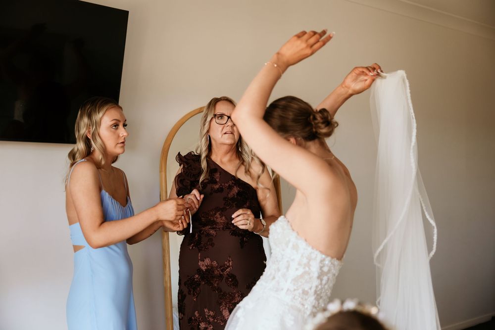 Two women help a bride attach her veil while standing in front of an arched mirror indoors.
