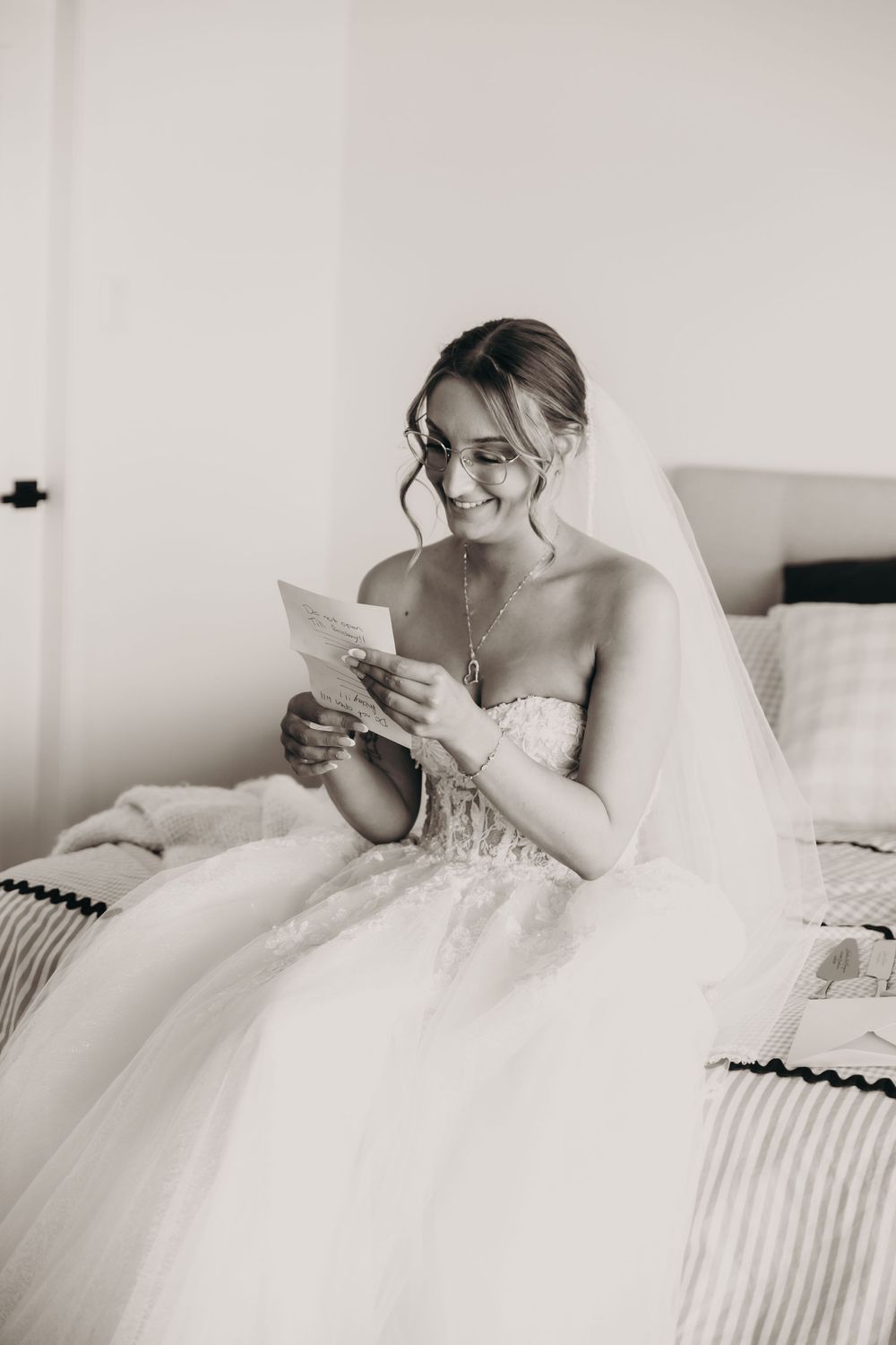 Smiling bride in strapless lace wedding gown reads a letter while seated on a bed.
