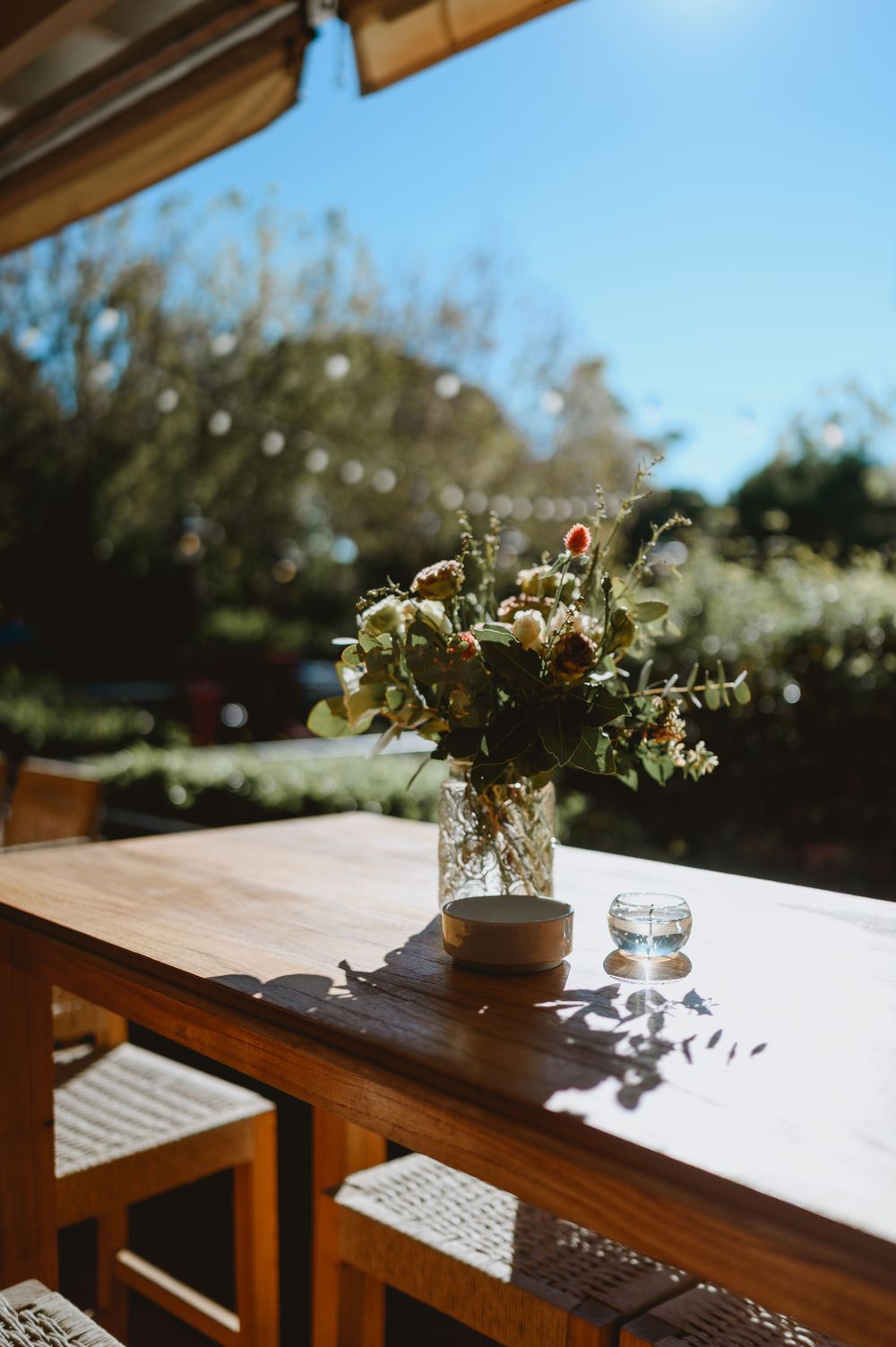 Rustic wooden table with a wildflower bouquet in a glass jar, set on a sunny outdoor patio with blue sky.