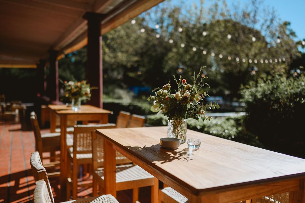 Sunlit outdoor patio with wooden tables decorated with wildflower bouquets and string lights in background.