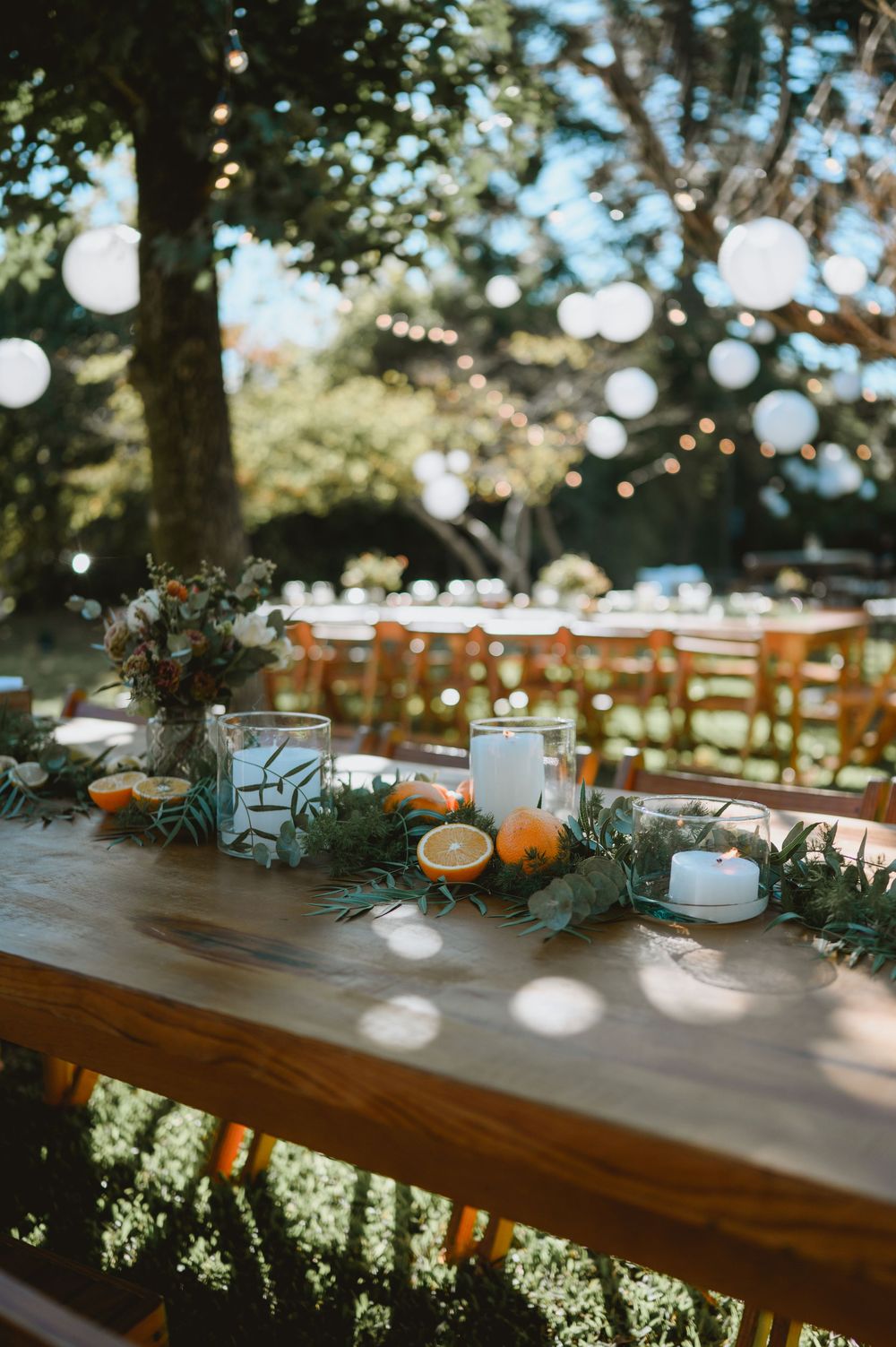 Wedding table centerpiece with candles, oranges, greenery garland, and globe string lights overhead.