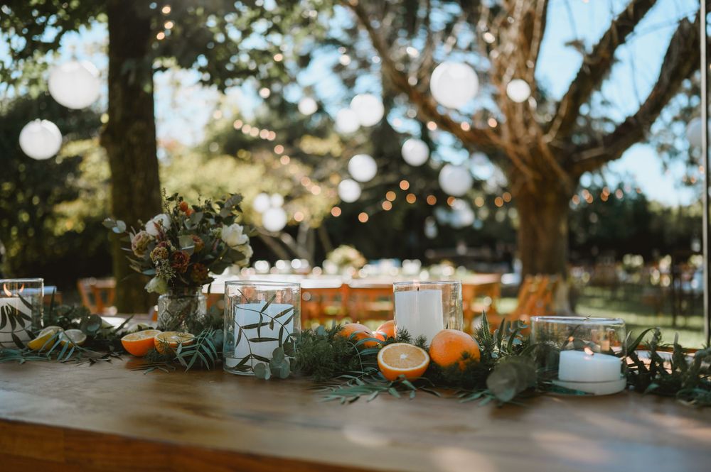 Outdoor wedding reception table with orange fruit, candles, greenery garland, and bokeh string lights.
