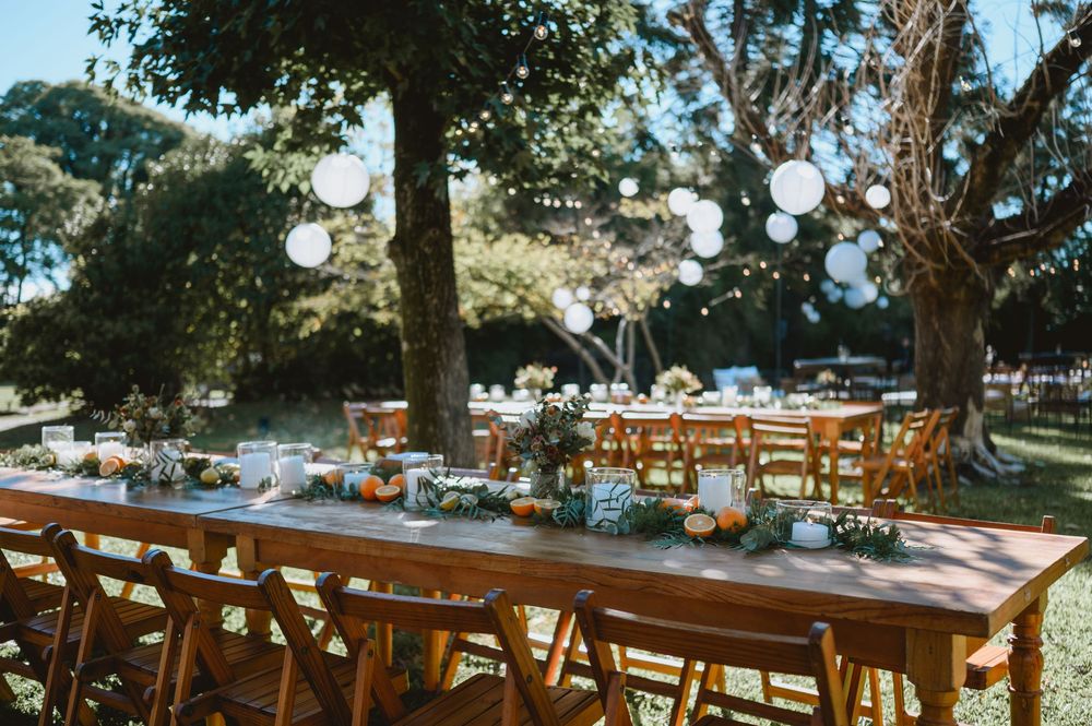 Elegant outdoor wedding reception with long wooden tables, white paper lanterns hanging from trees, and floral centerpieces.