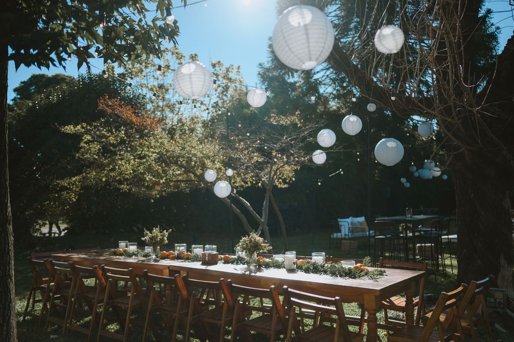 Romantic outdoor wedding dinner setup with wooden tables under trees adorned with white paper lanterns in natural light.