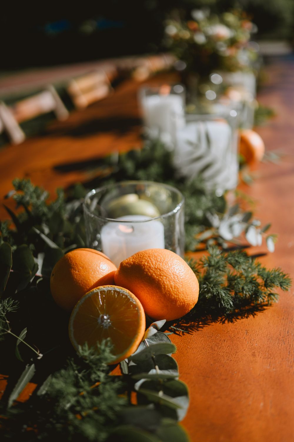 Close-up of rustic wedding table decor with fresh oranges, greenery garland, and glowing candles in glass holders.