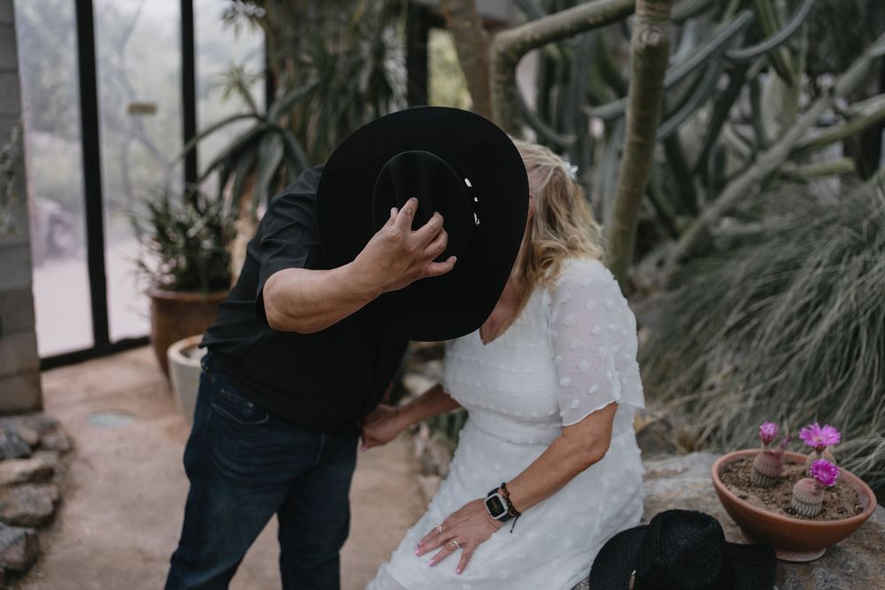Couple sharing a romantic kiss hidden behind a large black cowboy hat in a desert garden setting at Albuquerque Biopark