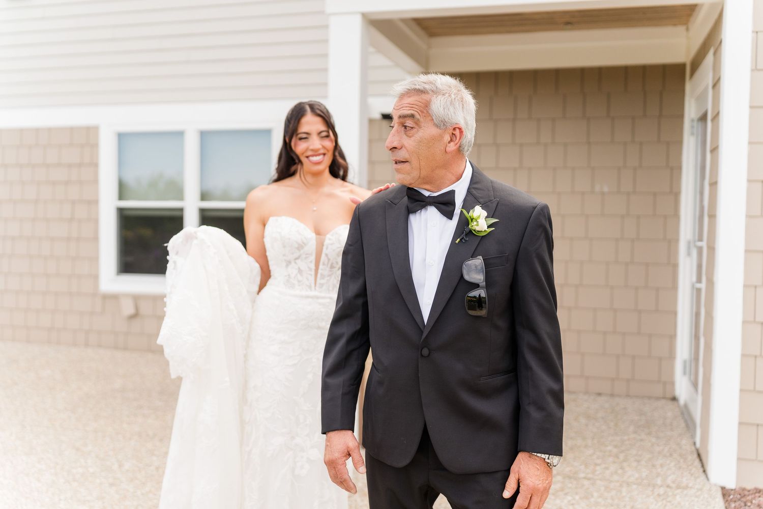 Bride in white lace gown smiles as father in tuxedo turns for first look outside.