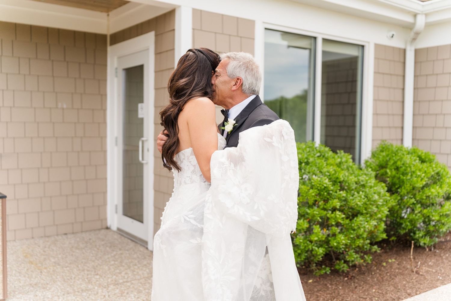 Bride in lace wedding gown shares a tender kiss with older man in tuxedo outside a building with green shrubs.