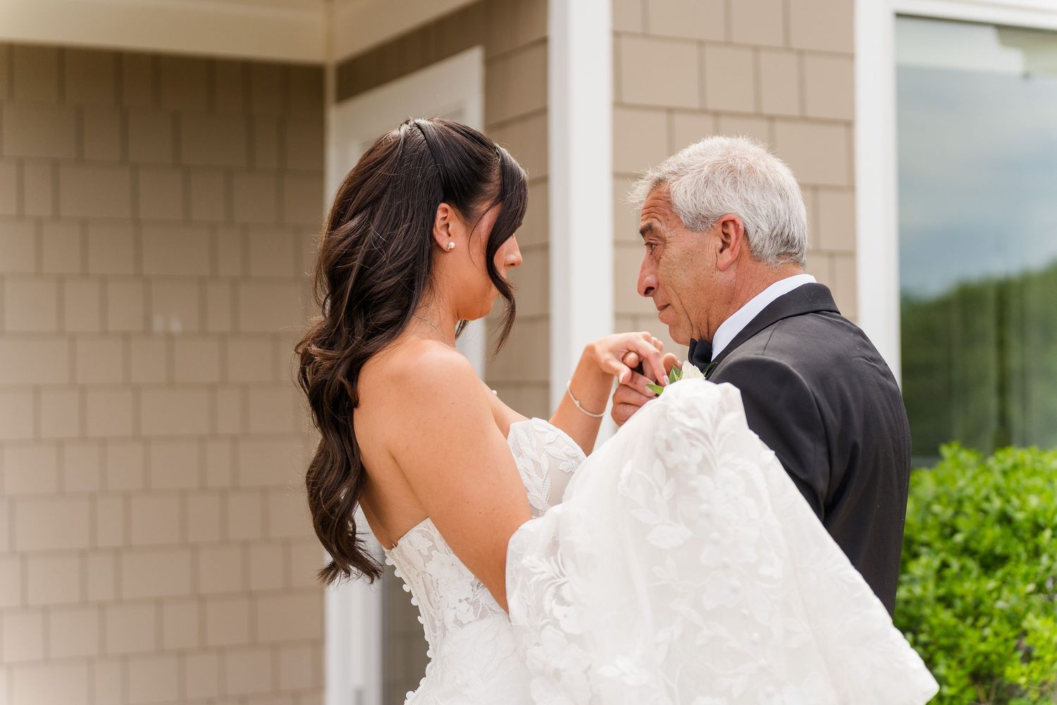 Bride in white lace gown shares a tender moment with older man in suit outside a building on wedding day.