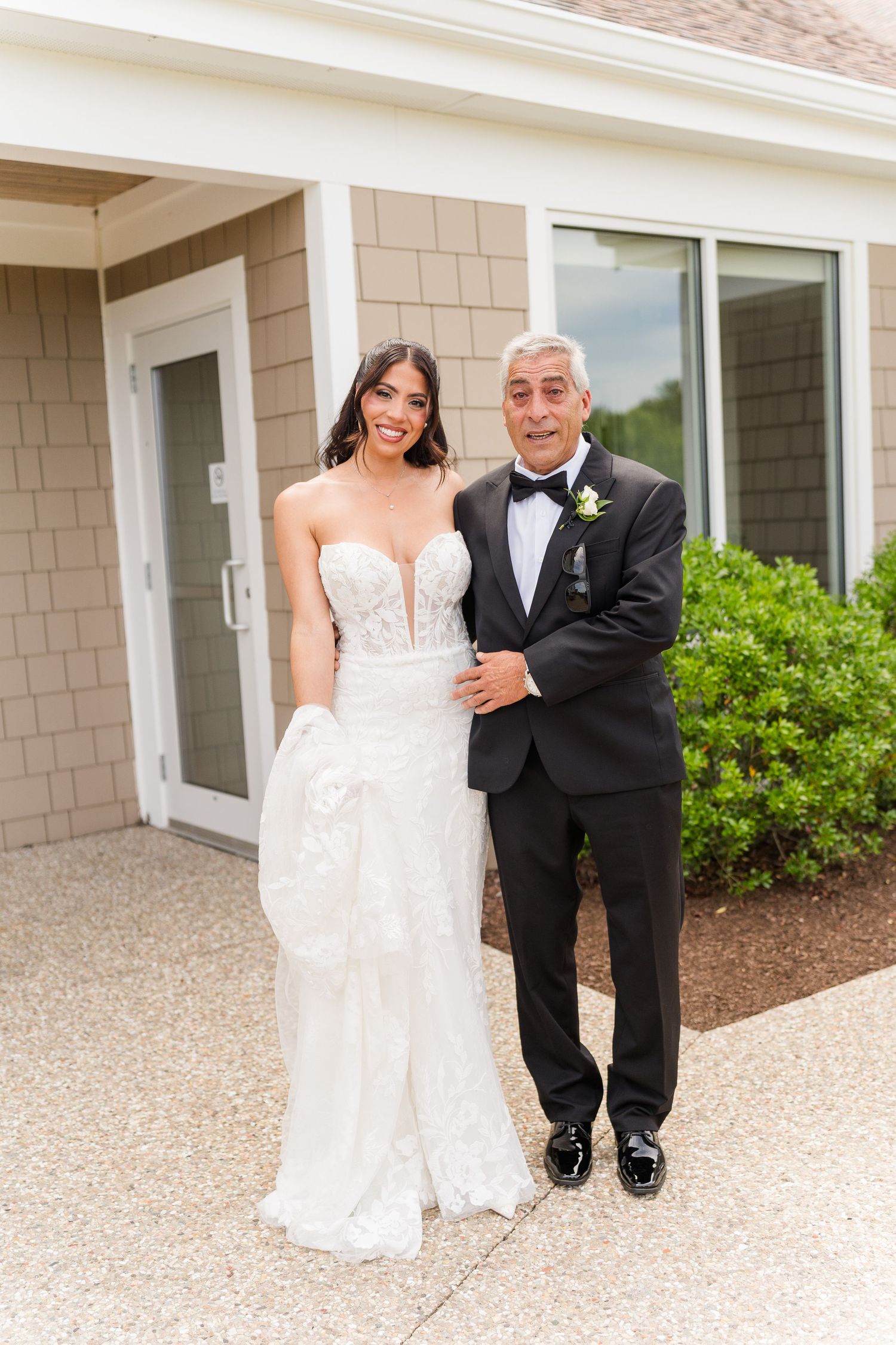 Bride in white lace gown and groom in black tuxedo posing together outside a building on their wedding day.