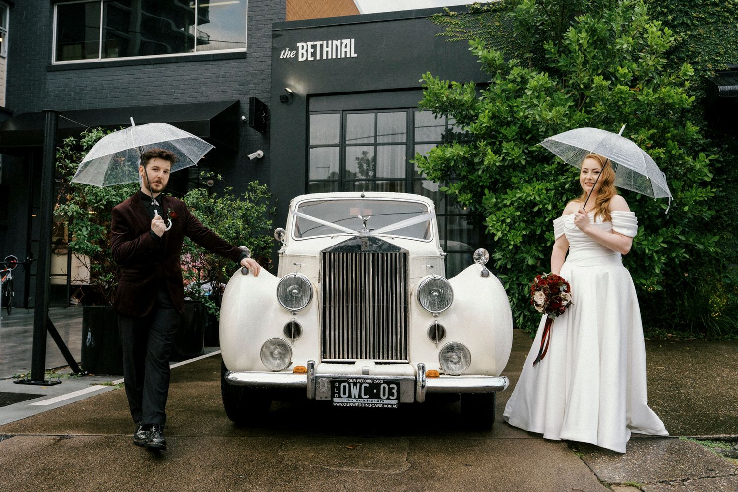 Bride and groom holding umbrellas beside a vintage white Rolls-Royce on a rainy wedding day outside the bethnal. Newstead Brisbane