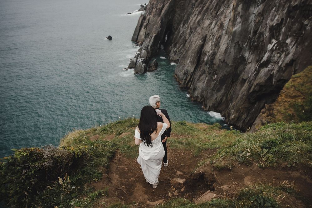 Couple in white attire standing on a dramatic coastal cliff edge overlooking turquoise ocean waters.