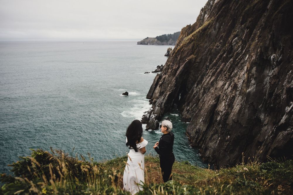 Couple holding hands on a dramatic coastal cliff edge with rocky ocean views on a misty overcast day.