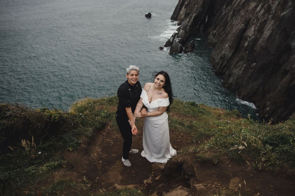 Couple poses on a dramatic coastal cliff edge in Ireland, woman in white dress, man in black, with turquoise sea below.