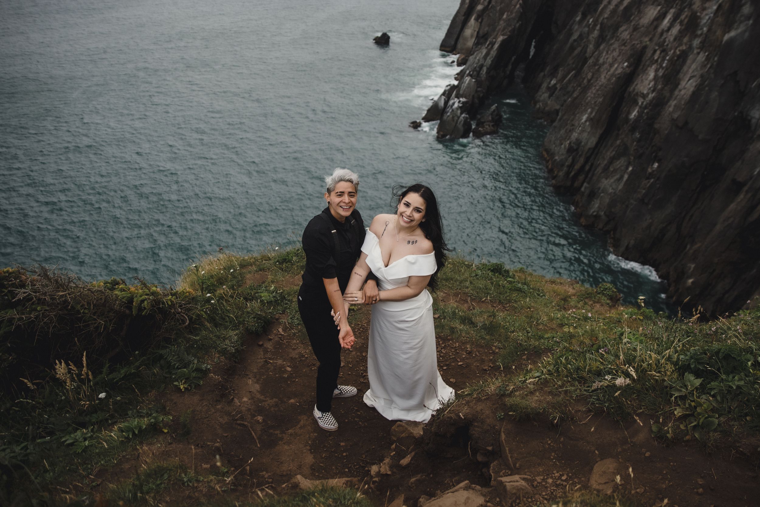 Couple poses on a dramatic coastal cliff edge in Ireland, woman in white dress, man in black, with turquoise sea below.