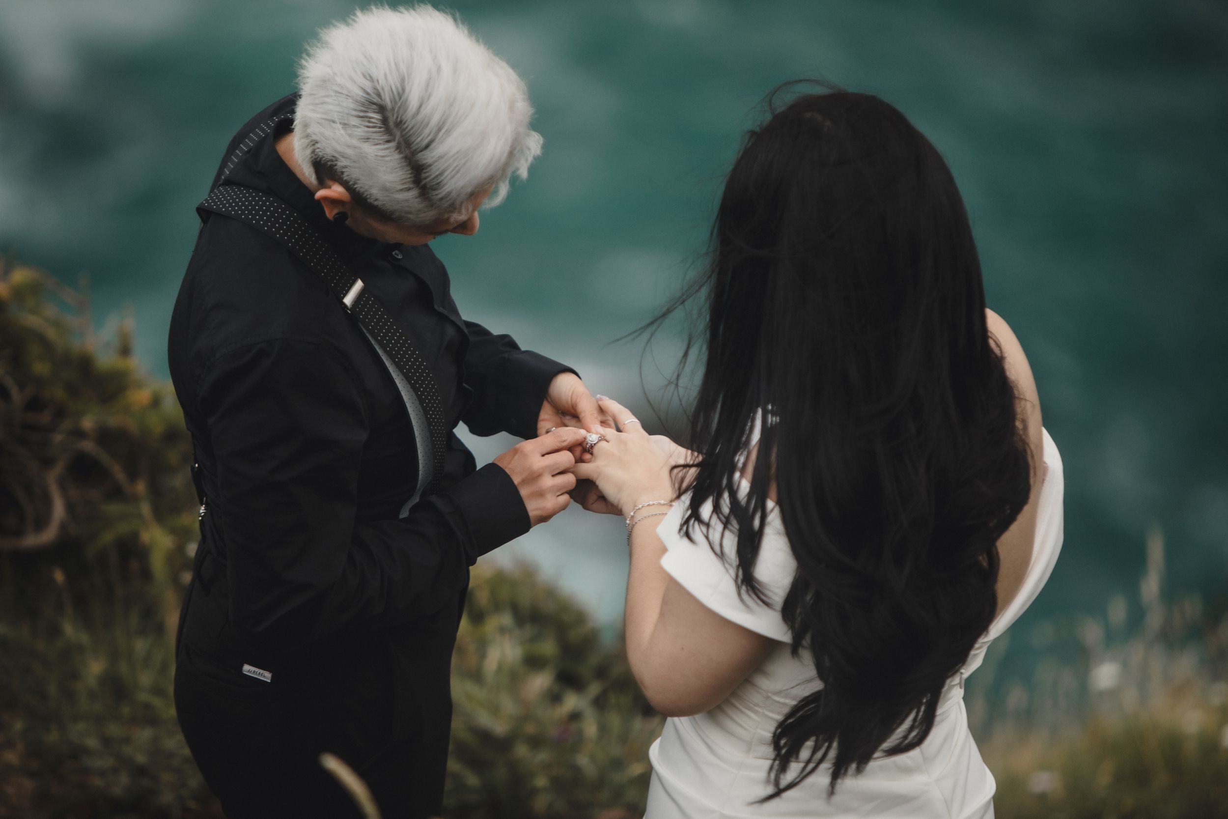 Person with white hair placing a ring on a dark-haired woman's finger outdoors with a scenic green background.