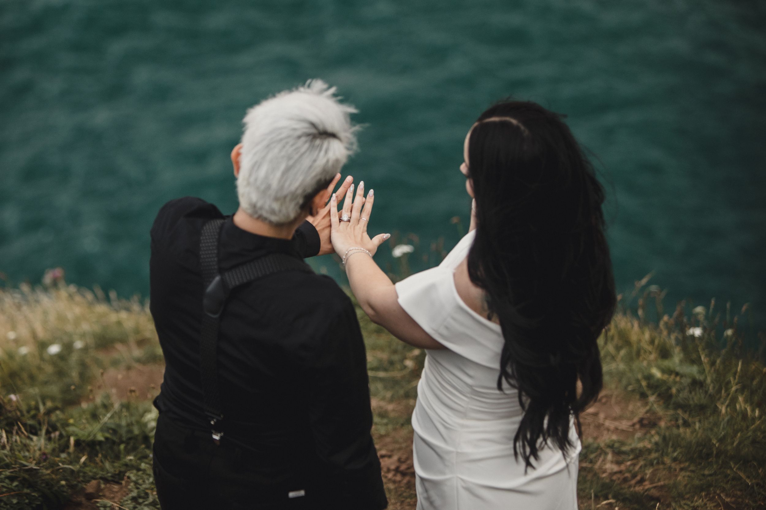 Two women, one with short gray hair and one with long dark hair, sit together overlooking a turquoise lake.