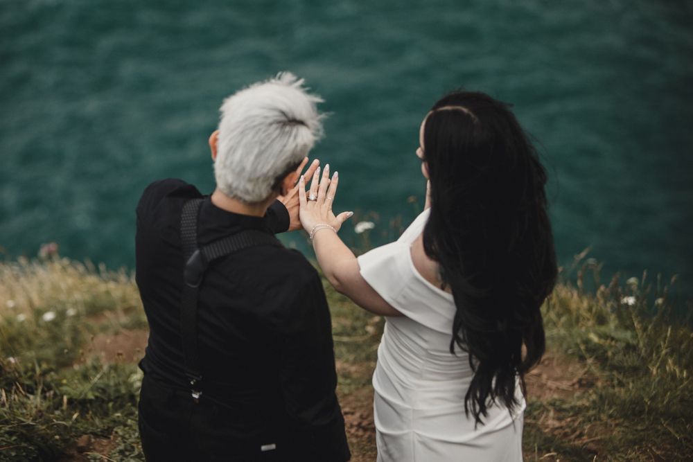 Two women, one with short gray hair and one with long dark hair, sit together overlooking a turquoise lake.
