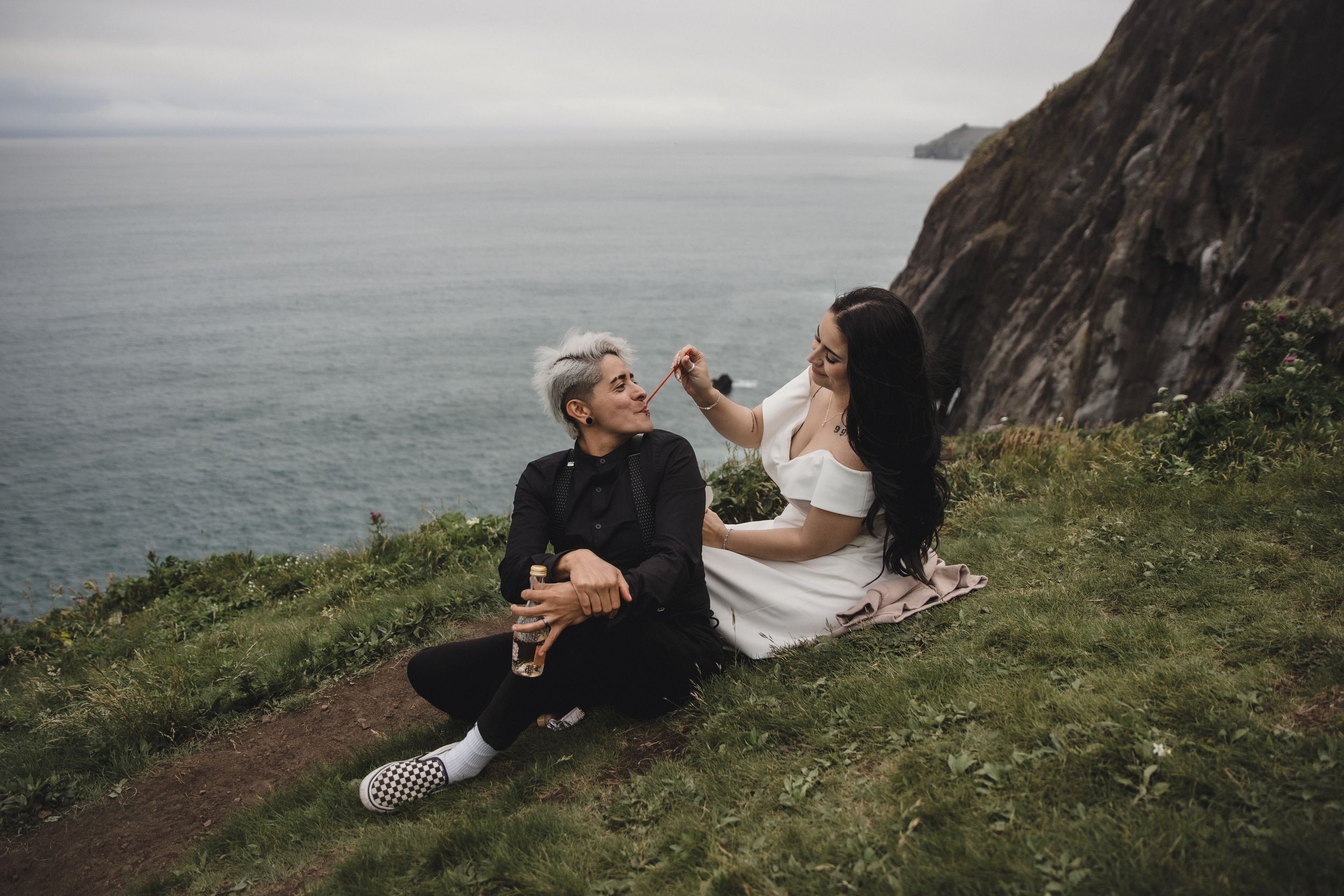 Couple sitting on cliffside grass by the ocean, sharing a romantic moment during their elopement shoot.