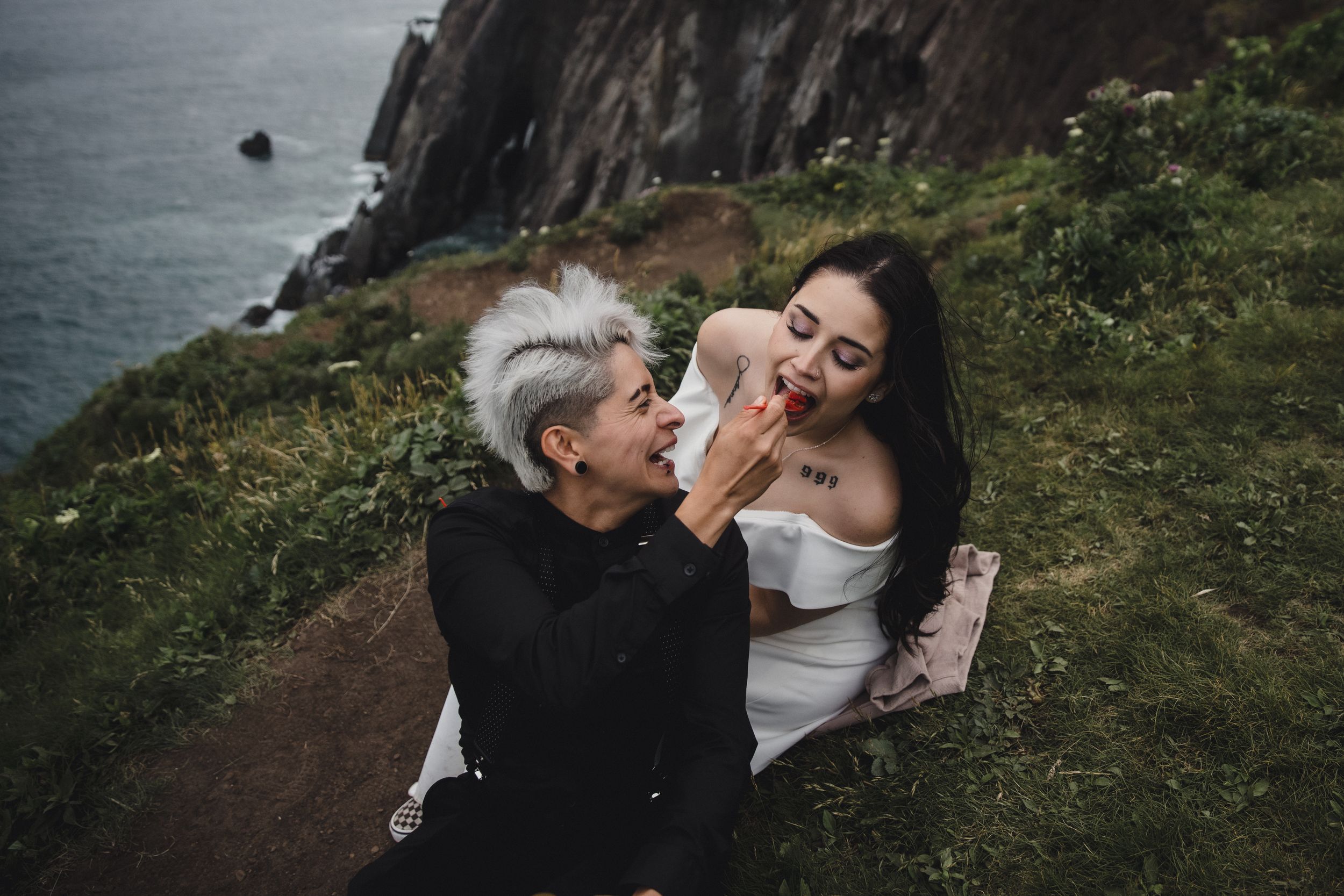 Two women share a romantic moment on a scenic coastal cliff, one feeding the other while dressed in elegant attire.