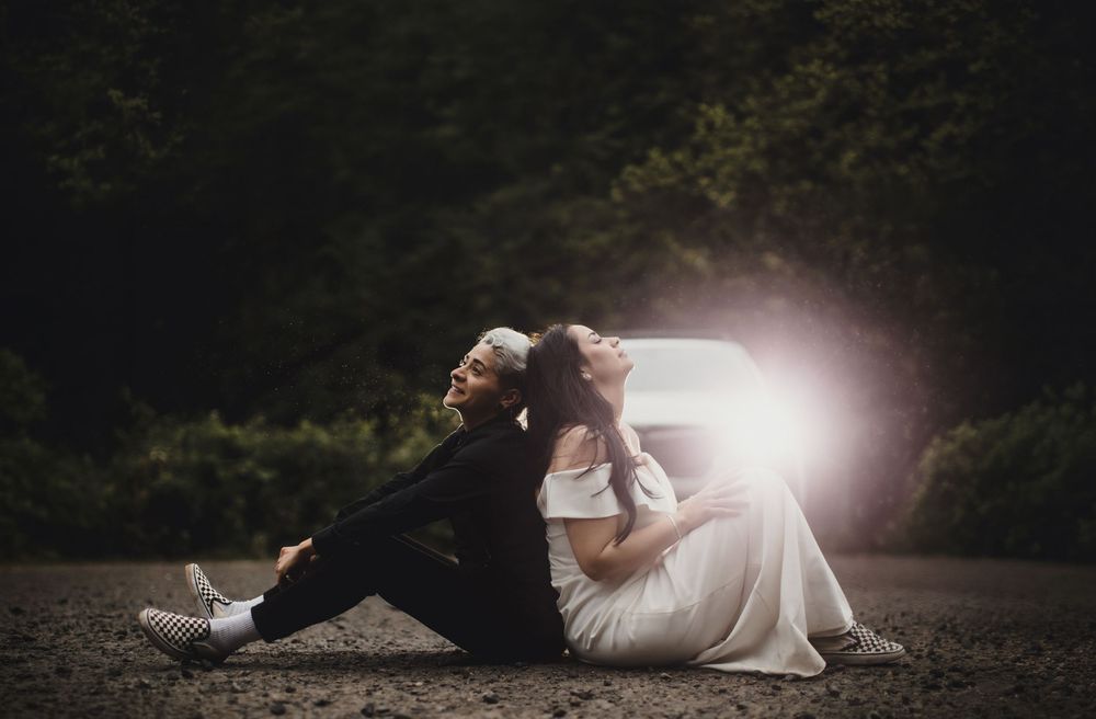 Two newlyweds sit back-to-back on a gravel path surrounded by dark trees, bathed in a soft glowing light.