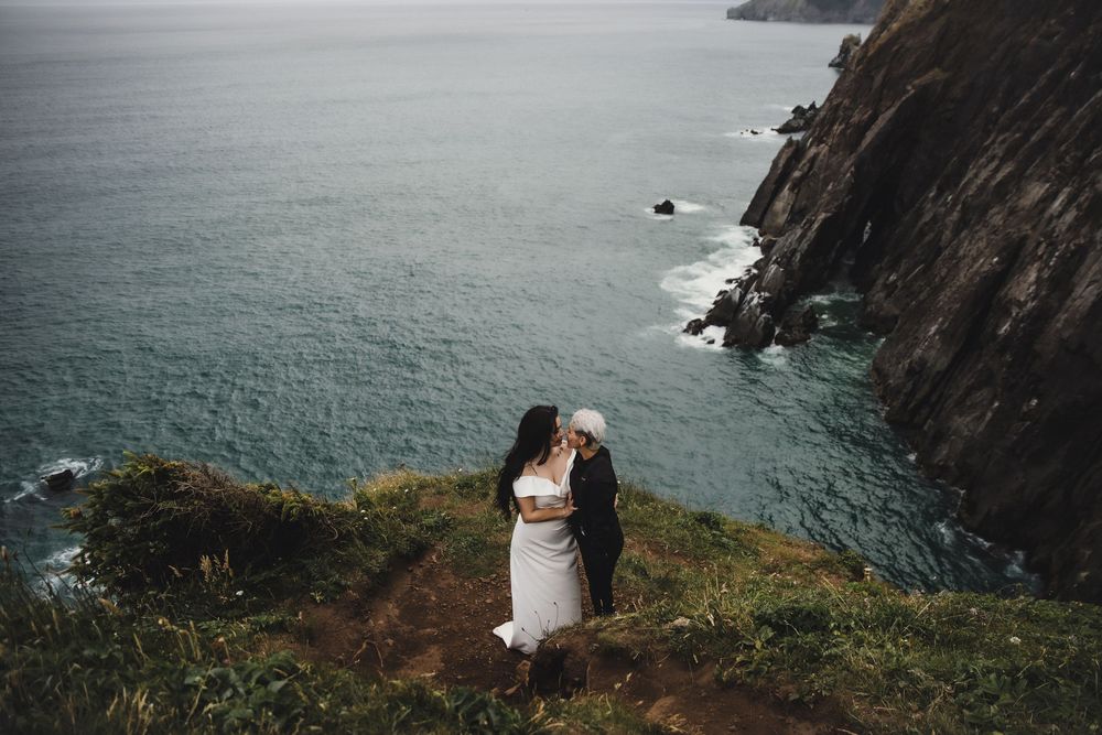 A couple shares a romantic embrace on a dramatic coastal cliff overlooking turquoise ocean waters.