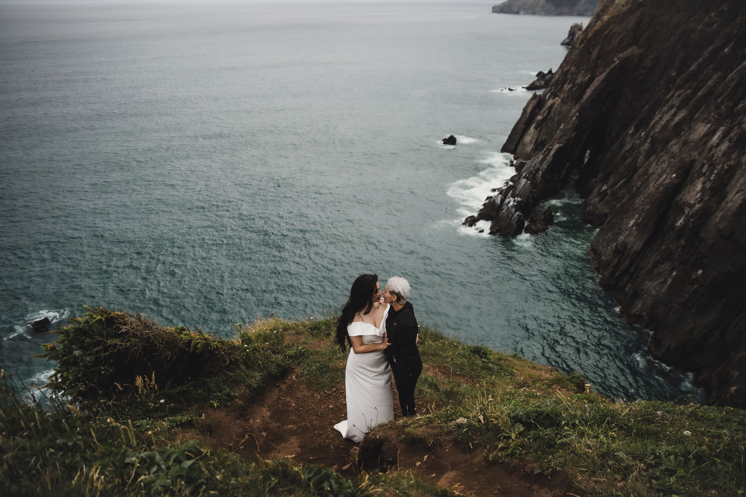 A couple shares a romantic embrace on a dramatic coastal cliff overlooking turquoise ocean waters.