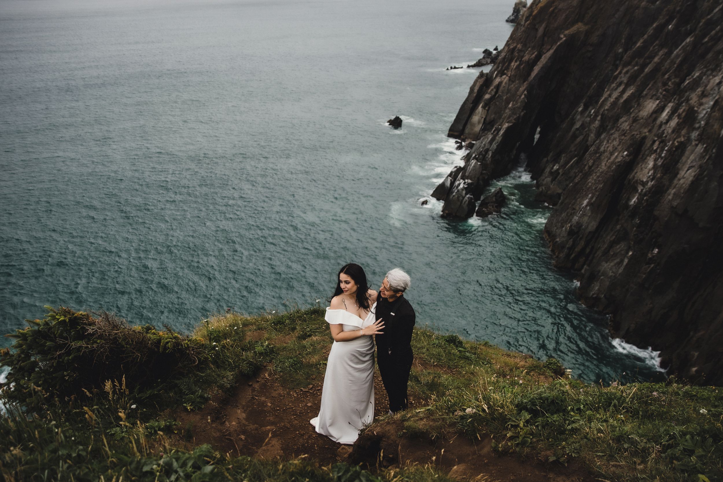 Couple embracing on a dramatic coastal cliff edge with turquoise ocean and rocky cliffs behind them.
