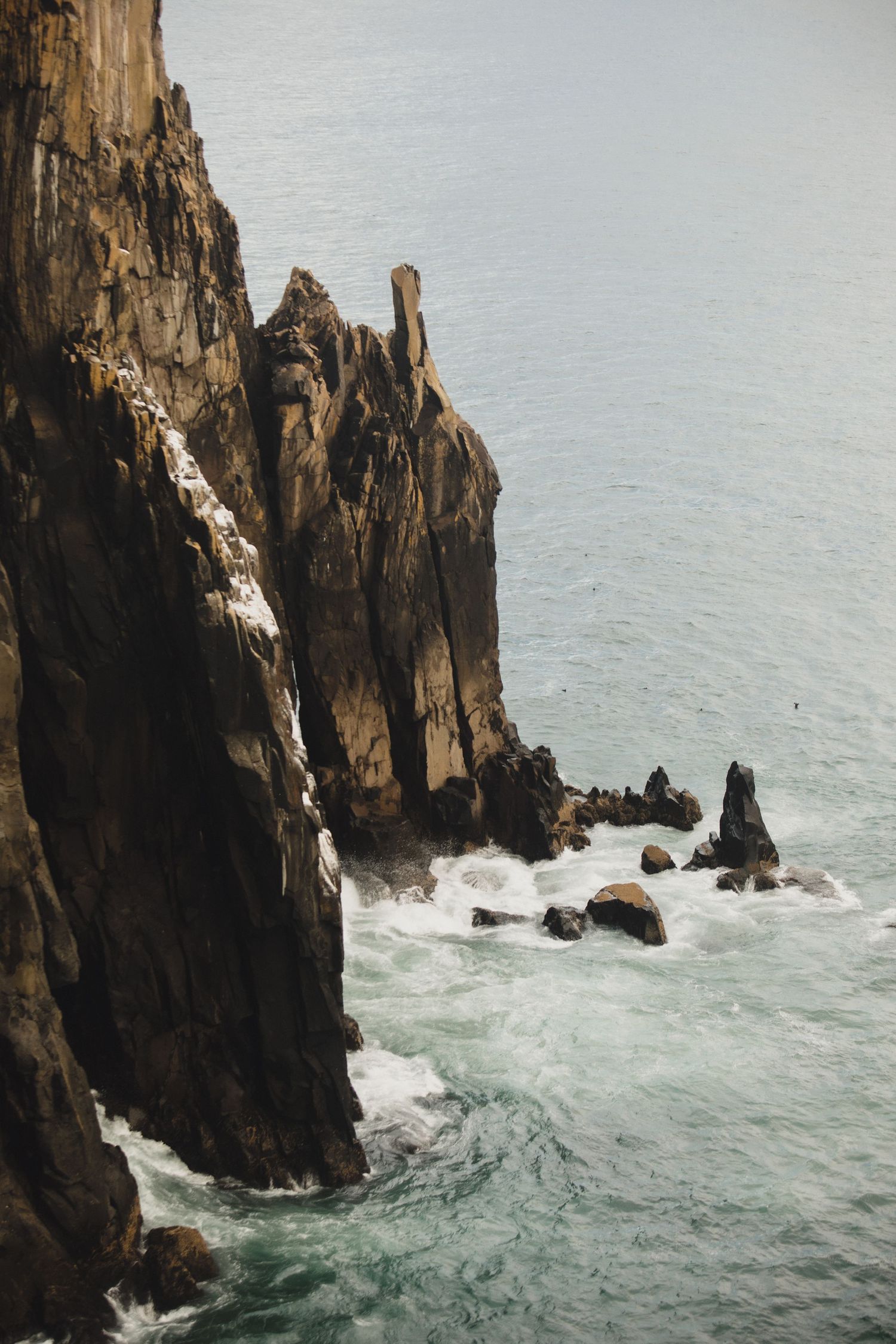 Dramatic coastal cliffs with green summit drop sharply to churning turquoise ocean waves below under overcast sky.