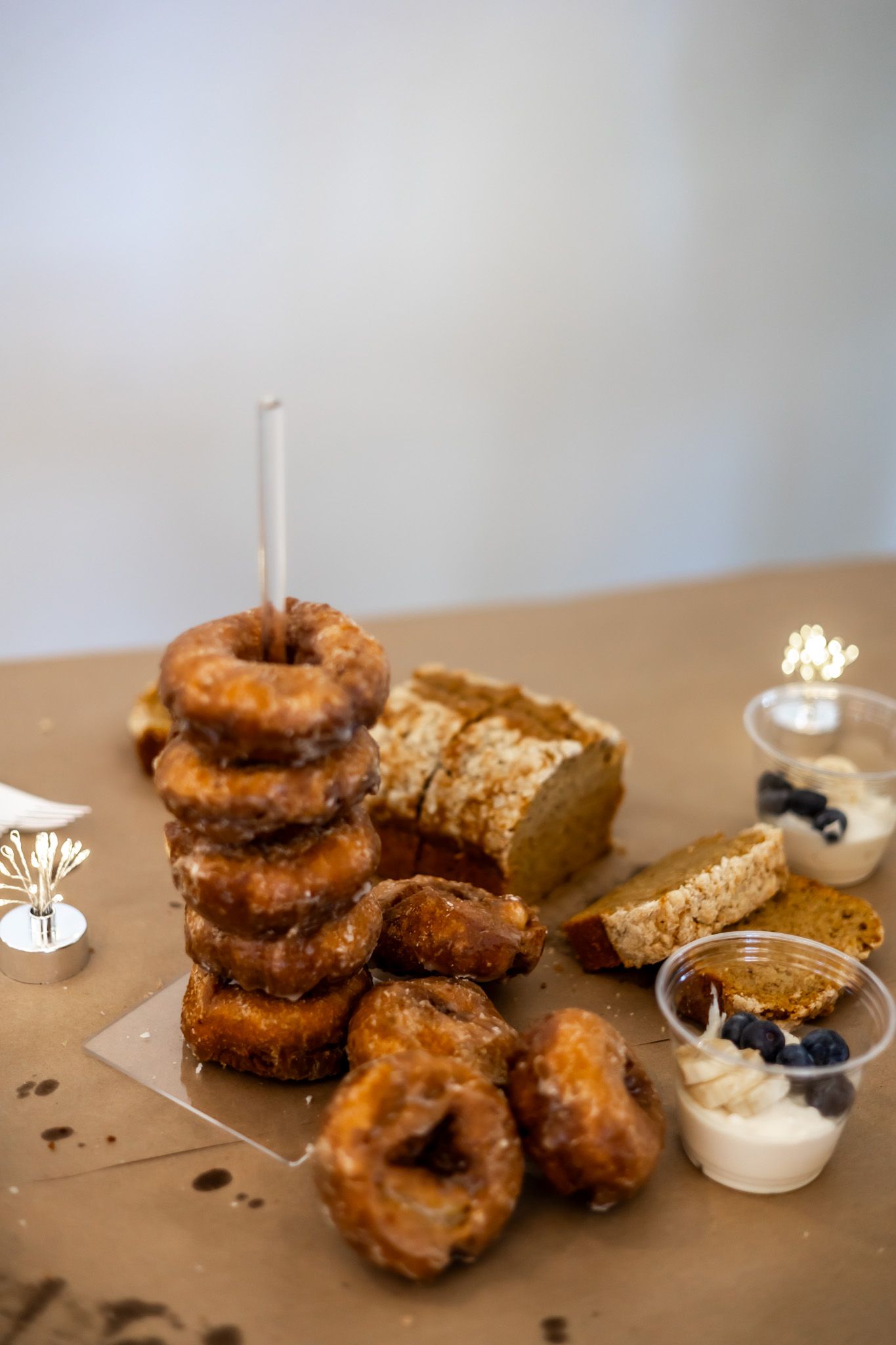 Stack of glazed donuts with a candle on a table beside bread slices, blueberries, and cream in small cups.
