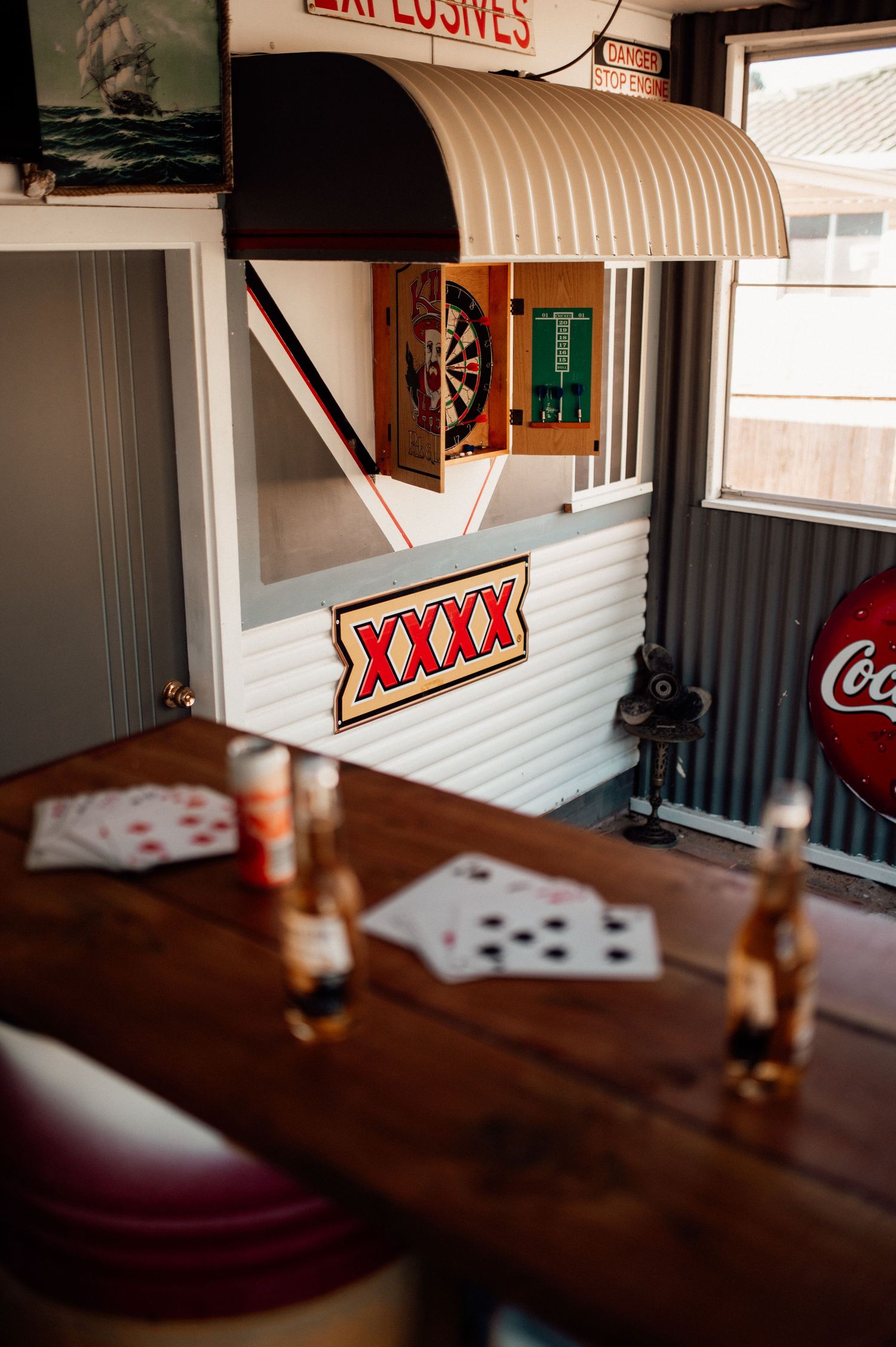 A rustic bar area with XXXX beer sign, dartboard, Coca-Cola sign, playing cards and beer bottles on a wooden table.