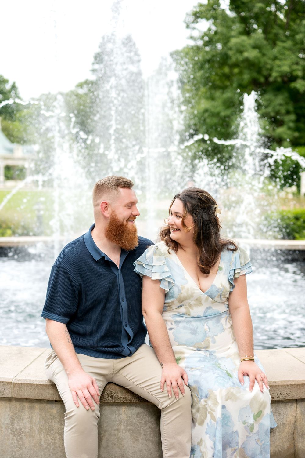 A smiling couple sits together on a fountain ledge outdoors, the man in navy and the woman in a floral dress.