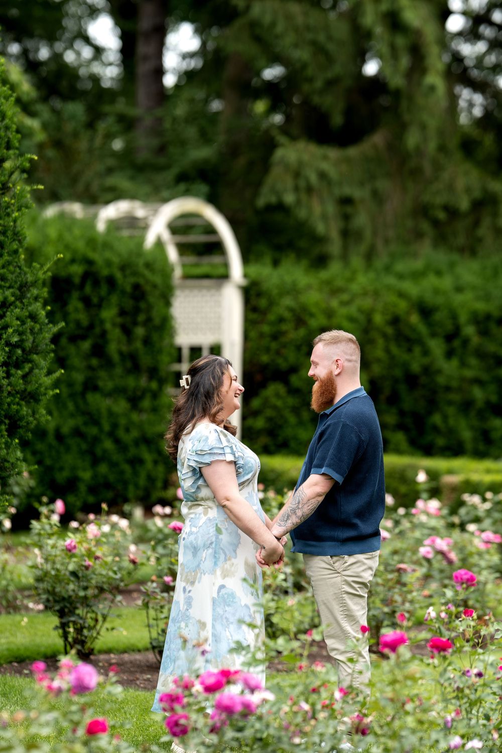 Couple holding hands and smiling at each other in a blooming rose garden with a white arbor backdrop.